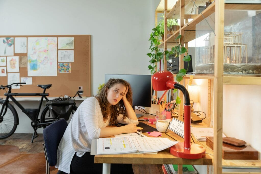 Tired businesswoman working at a cluttered desk, surrounded by office supplies and plants.